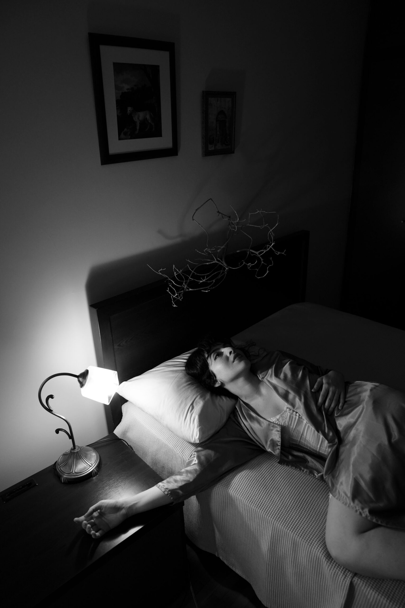 Mulher deitada de lado na cama à noite, com o braço estendido sobre a cômoda, em cena que remete ao instante entre o sono e a paralisia do sono