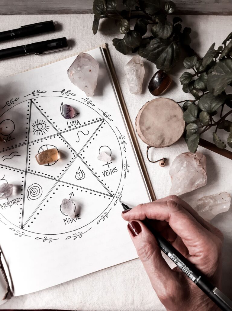 Mão desenhando pentagrama com símbolos planetários — Sol, Lua, Vênus, Marte e Mercúrio — em caderno esotérico com cristais e pedras ao redor