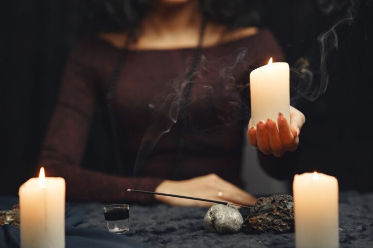 Mulher segurando vela acesa em ritual mágico com velas, incenso, pedras e recipiente sobre mesa escura