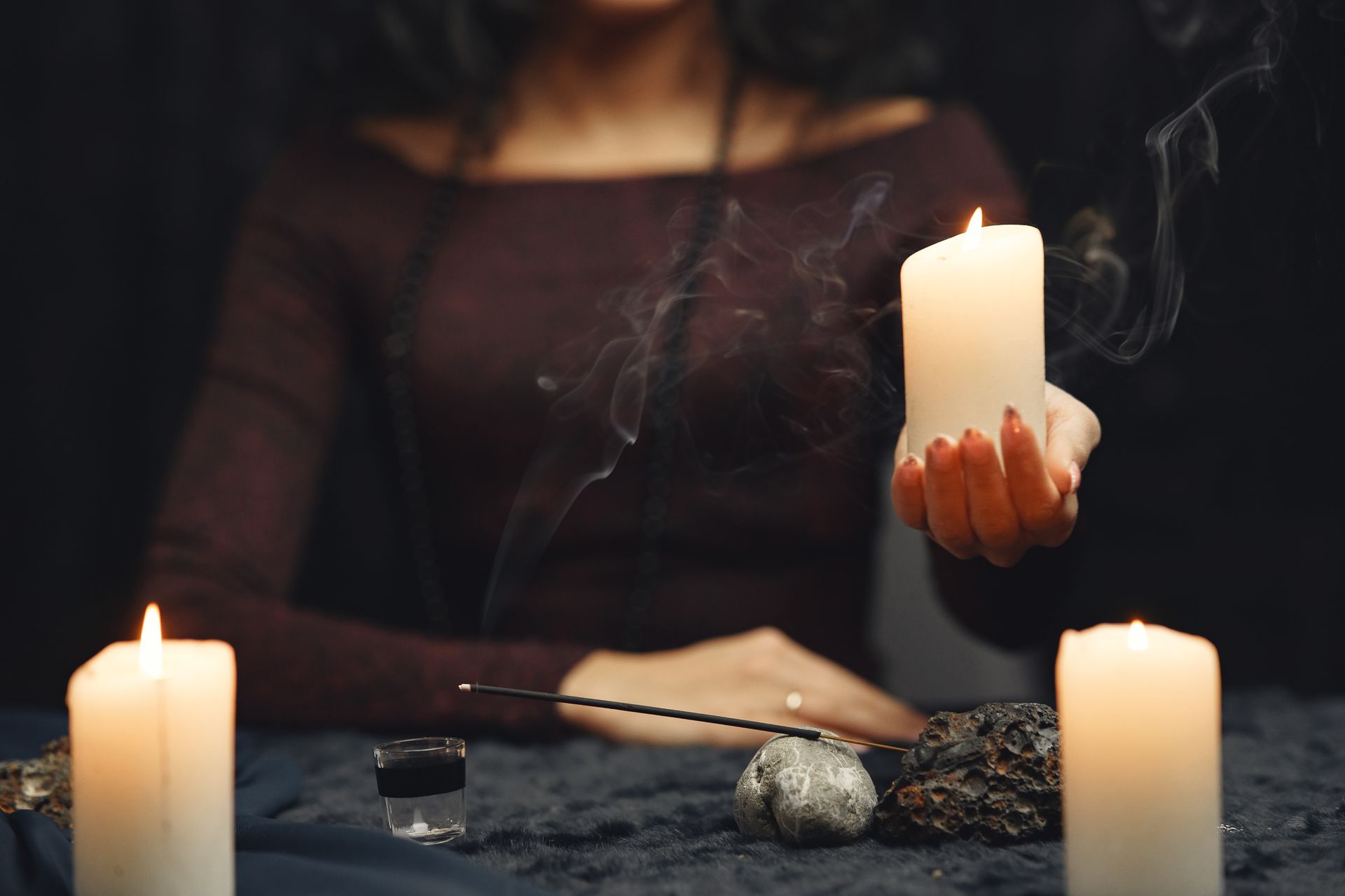 Mulher segurando vela acesa em ritual mágico com velas, incenso, pedras e recipiente sobre mesa escura
