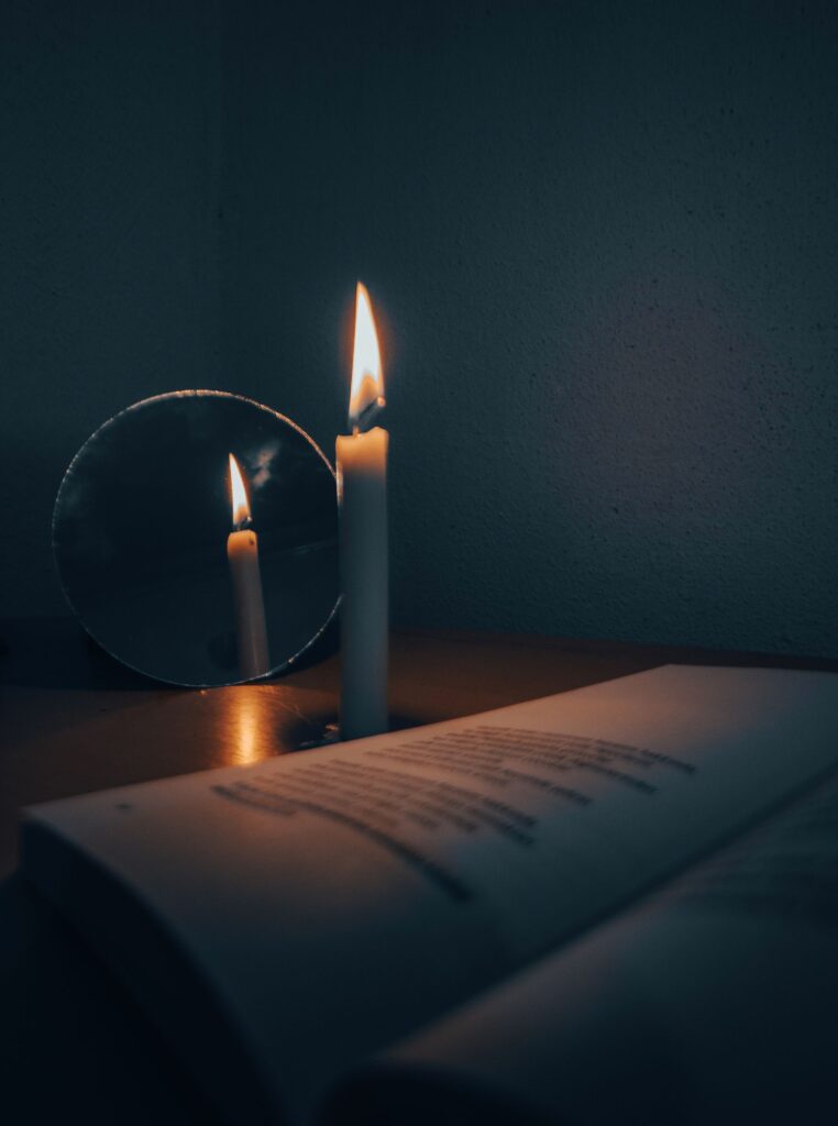 Vela acesa refletida em um pequeno espelho ao lado de um livro aberto, em ambiente escuro, simbolizando o grimório como espelho da mente