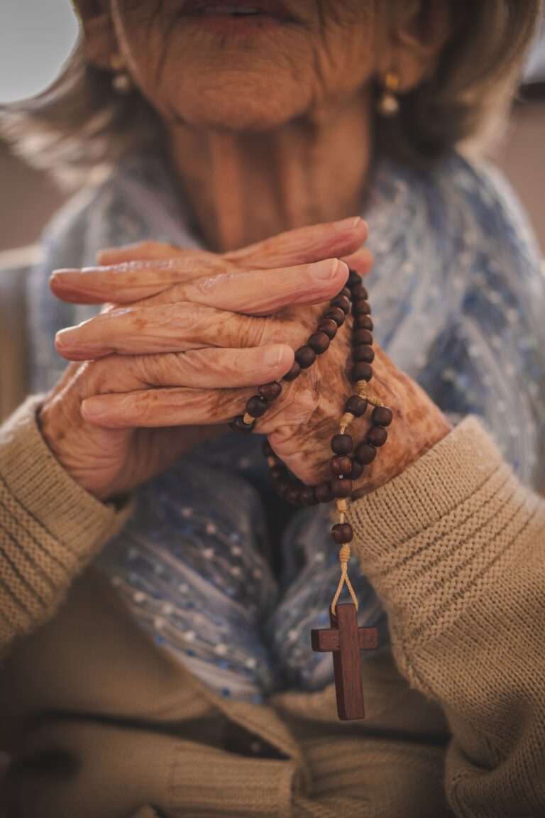 Mãos idosas em oração segurando um terço de madeira com crucifixo, representando a fé e a tradição das benzedeiras brasileiras