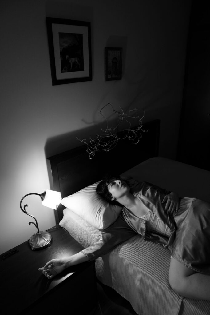 Mulher deitada de lado na cama à noite, com o braço estendido sobre a cômoda, em cena que remete ao instante entre o sono e a paralisia do sono