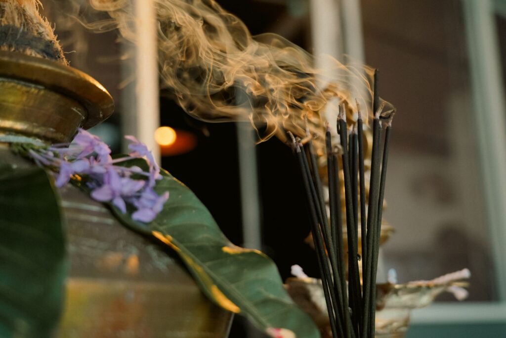 Varetas de incenso queimando com fumaça em altar ritual, ao lado de flor lilás e vaso de bronze