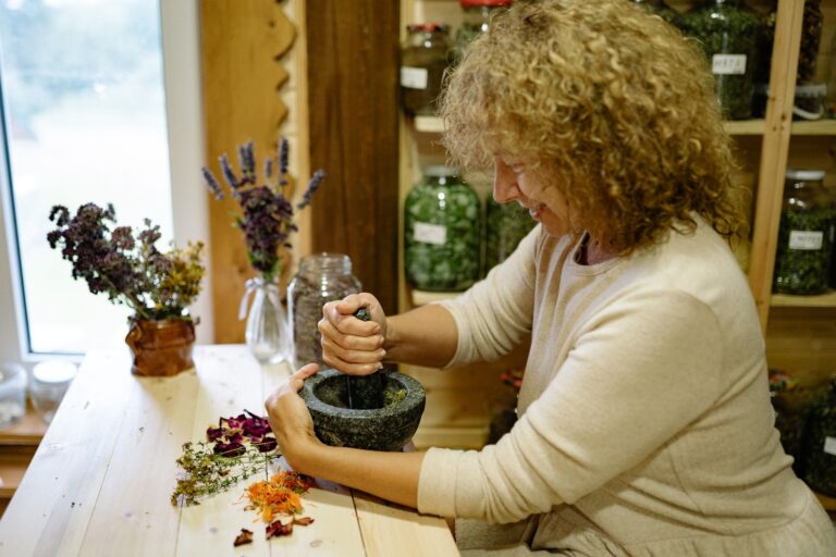 Mulher preparando ervas medicinais em pilão de pedra, cercada por potes de vidro com plantas secas e flores sobre a mesa