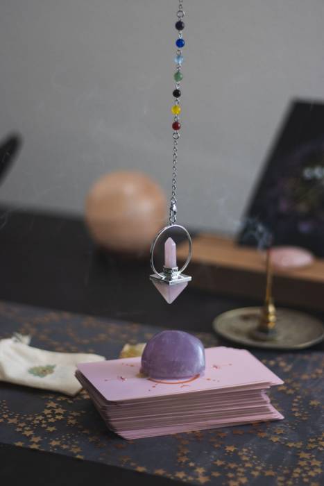 Pêndulo de radiestesia com ponta de quartzo rosa e corrente com contas coloridas dos chakras, pousado sobre um baralho de tarot rosa ao lado de uma pedra de ametista